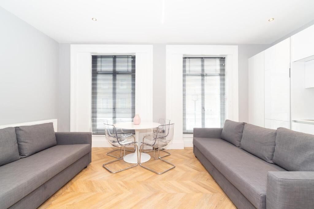 Una sala de estar moderna con un sofá gris, una mesa redonda con sillas y grandes ventanales con persianas.