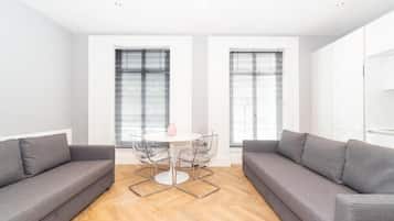 Una sala de estar moderna con un sofá gris, una mesa redonda con sillas y grandes ventanales con persianas.