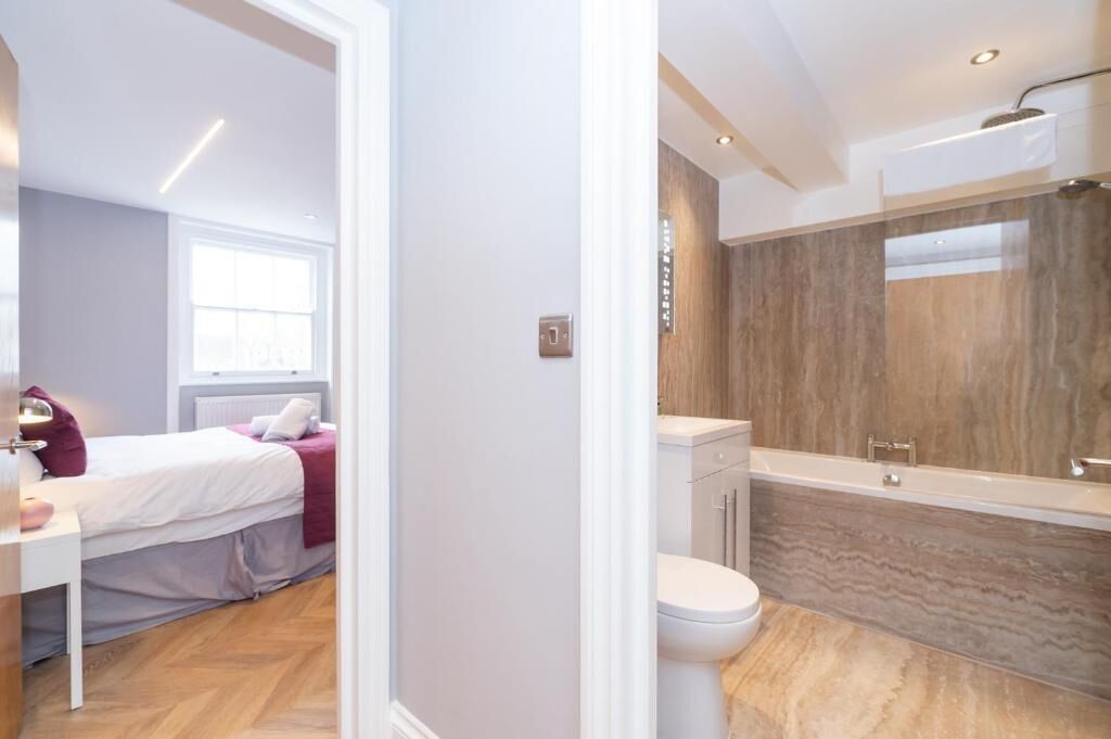 Un dormitorio moderno con cama, mesita de noche y ventana. Junto a él, un baño con bañera, inodoro y un espejo grande.