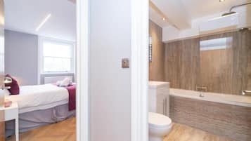 Un dormitorio moderno con cama, mesita de noche y ventana. Junto a él, un baño con bañera, inodoro y un espejo grande.
