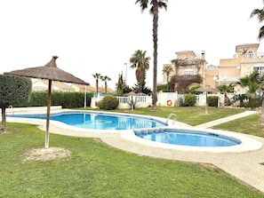 A swimming pool with a thatched umbrella, surrounded by well-maintained grass and palm trees. - Detached 3-Bed Villa with Shared Pool near La Zenia, Orihuela Costa (Orihuela)