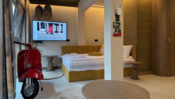 Ett modernt hotellrum med en säng, en röd skoter, en tv som visar en gatuscen och en garderob i trä.