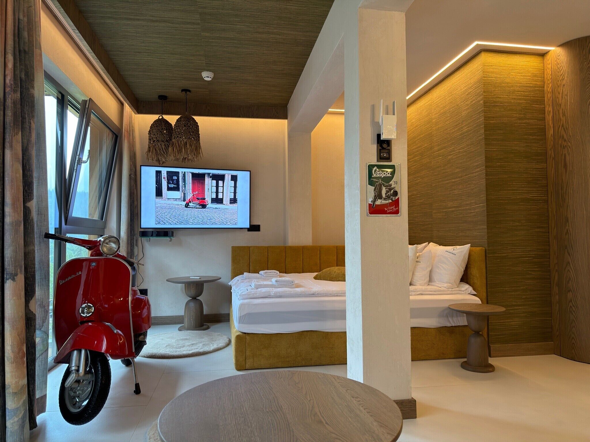 A hotel room with a bed, a round table, a red scooter, and a television mounted on the wall.
