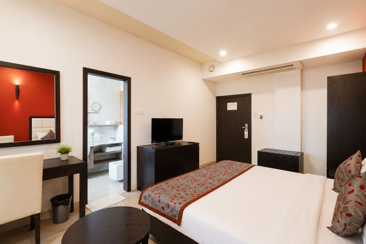 Habitación de hotel con cama, escritorio, televisión, espejo y baño visible a través de una puerta abierta.