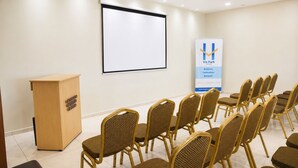 Un salón de conferencias con un atril, una pantalla de proyección y filas de sillas de mimbre.