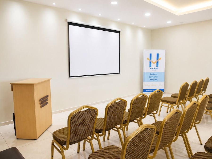 Uma sala de conferências com um púlpito, uma tela de projeção e fileiras de cadeiras de vime.
