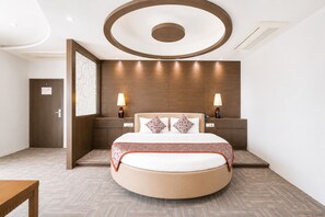 A modern hotel room with a round bed, wooden headboard, two bedside lamps, and a patterned throw blanket.