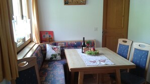 Een gezellig eetgedeelte met een houten tafel en blauwe fauteuils, een bank met gestreepte kussens, en een raam met gordijnen.