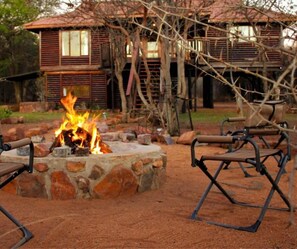 A rustic outdoor seating area with a stone fire pit and metal chairs, surrounded by a wooden deck and a house with a sloped roof. - Palala River Cottages (Waterberg)