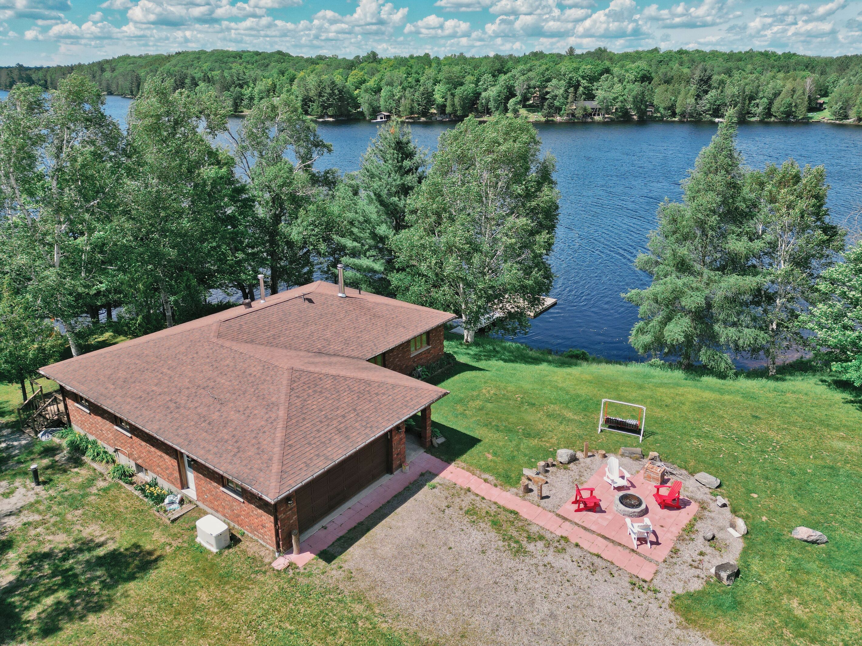 Un grand bâtiment avec un toit rouge, une terrasse dallée avec des sièges extérieurs, et une vue sur un lac entouré d’arbres.