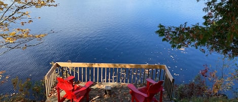 Ett trädäck med två röda Adirondack-stolar med utsikt över en stilla sjö.