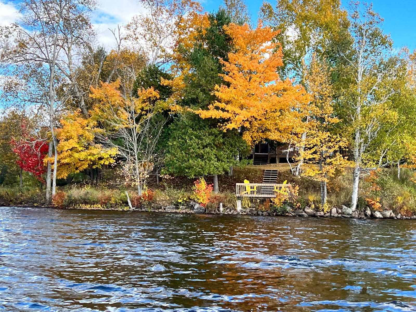 Une vue sur le lac, avec le feuillage automnal, une jetée en bois et une maison partiellement cachée par les arbres.