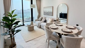 A modern living room with a dining area, a sofa, a coffee table, and a large window with a view. - 48 Parkside - 603 1BD (Dubai)