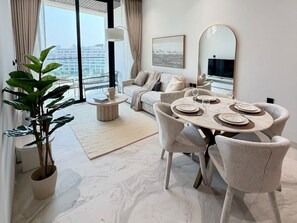 A modern living room with a dining area, a sofa, a coffee table, and a large window with a view. - 48 Parkside - 603 1BD (Dubai)