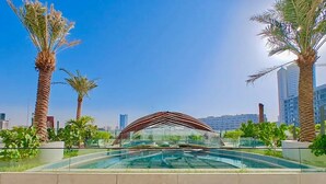 A swimming pool with a modern architectural canopy, surrounded by palm trees and urban buildings in the background. - 48 Parkside - 603 1BD (Dubai)