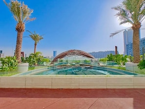 A swimming pool with a modern architectural canopy, surrounded by palm trees and urban buildings in the background. - 48 Parkside - 603 1BD (Dubai)