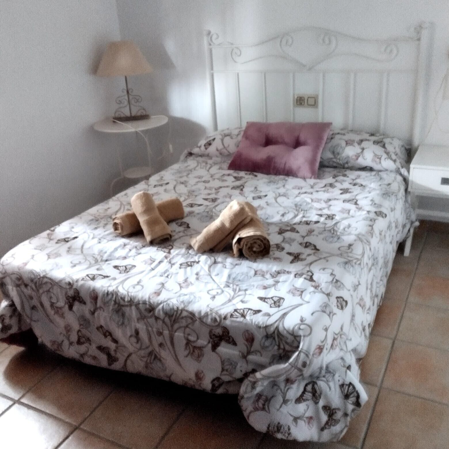 Una cama bien hecha con una colcha floral, una toalla enrollada y un cojín decorativo.