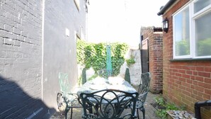 A patio with a table and chairs, a brick wall, and a window. - Cobblers Cottage (Reydon)