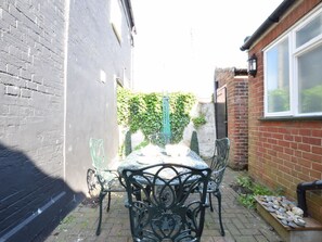 A patio with a table and chairs, a brick wall, and a window. - Cobblers Cottage (Reydon)