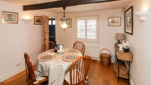 A dining area with a round table, wooden chairs, a hanging light fixture, and a window with blinds. - Cobblers Cottage (Reydon)