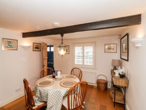 A dining area with a round table, wooden chairs, a hanging light fixture, and a window with blinds. - Cobblers Cottage (Reydon)