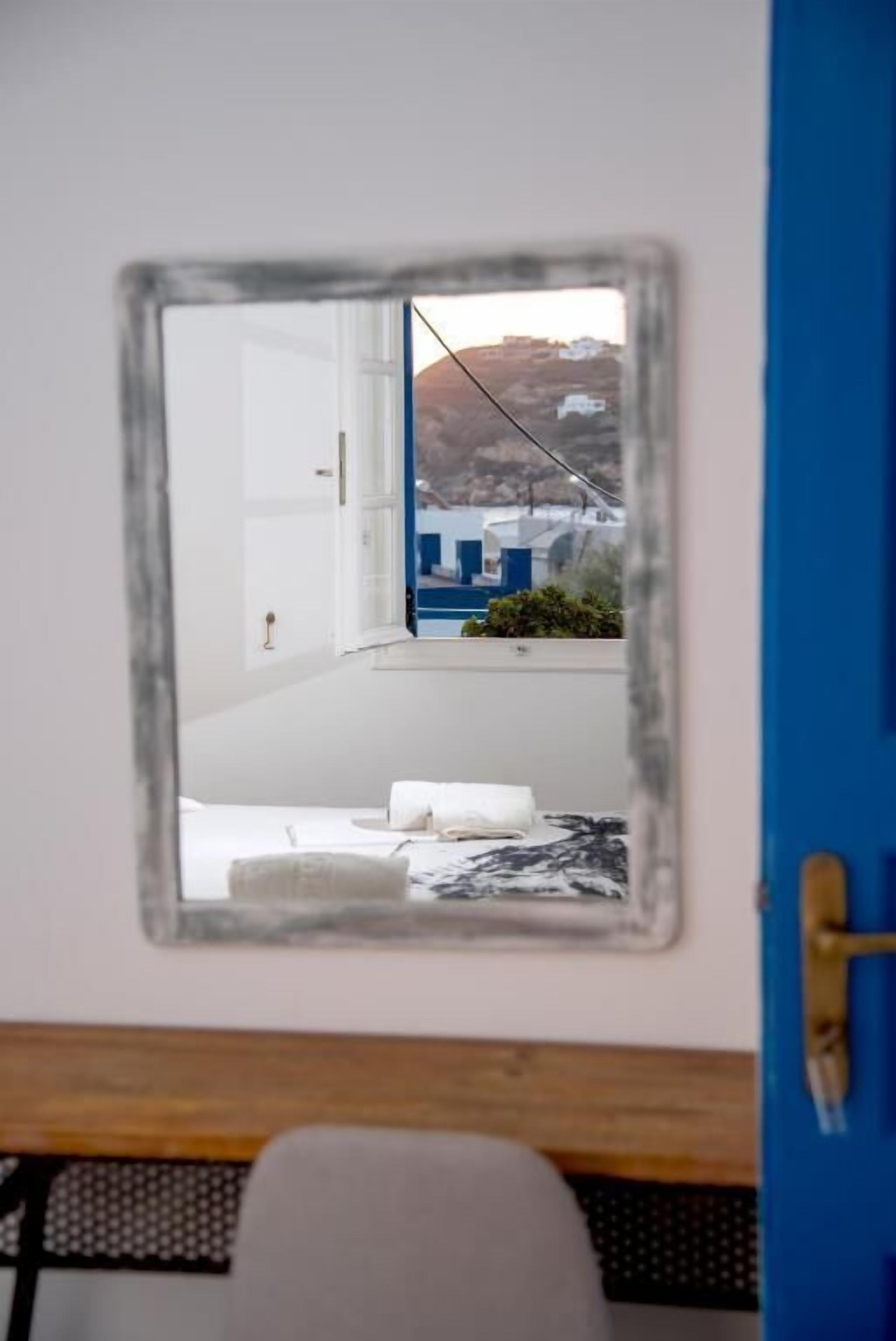 A mirror reflecting a room with a bed, a blue door, and a view of a building with white and blue structures.