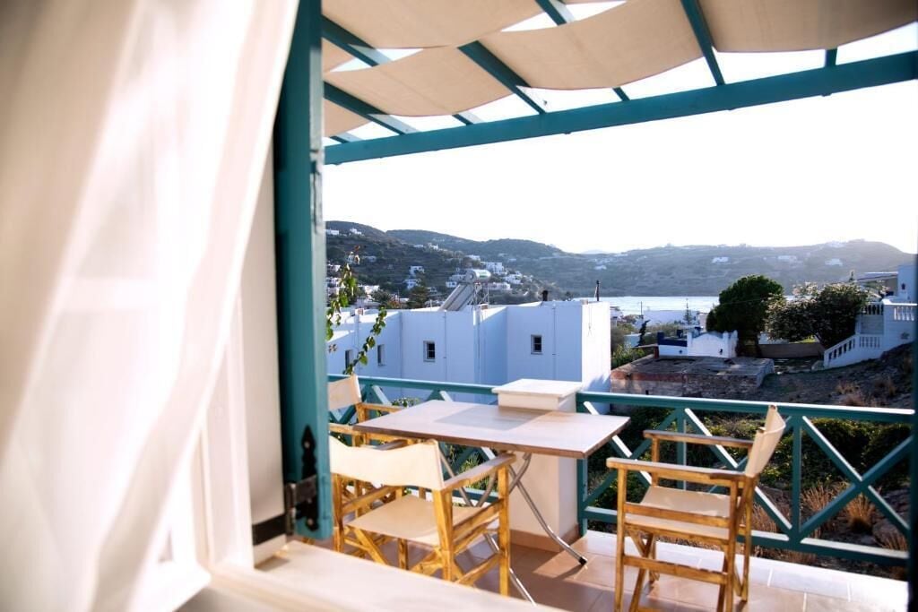A balcony with a table and chairs, overlooking a coastal town with buildings and hills.
