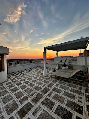 Eine Dachterrasse mit gemustertem Boden, einer überdachten Sitzgelegenheit und Blick auf den Sonnenuntergang.