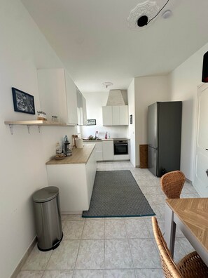 Eine moderne Küche mit weißen Schränken, einer Edelstahl-Mülltonne, einem Korbstuhl und einem Kühlschrank.