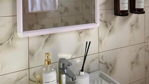 A bathroom with a marble-tiled wall, a white sink, a mirror with a pink frame, a towel hanging on the wall, and two bottles of hand soap. - Peakline Apartments (Arusha)
