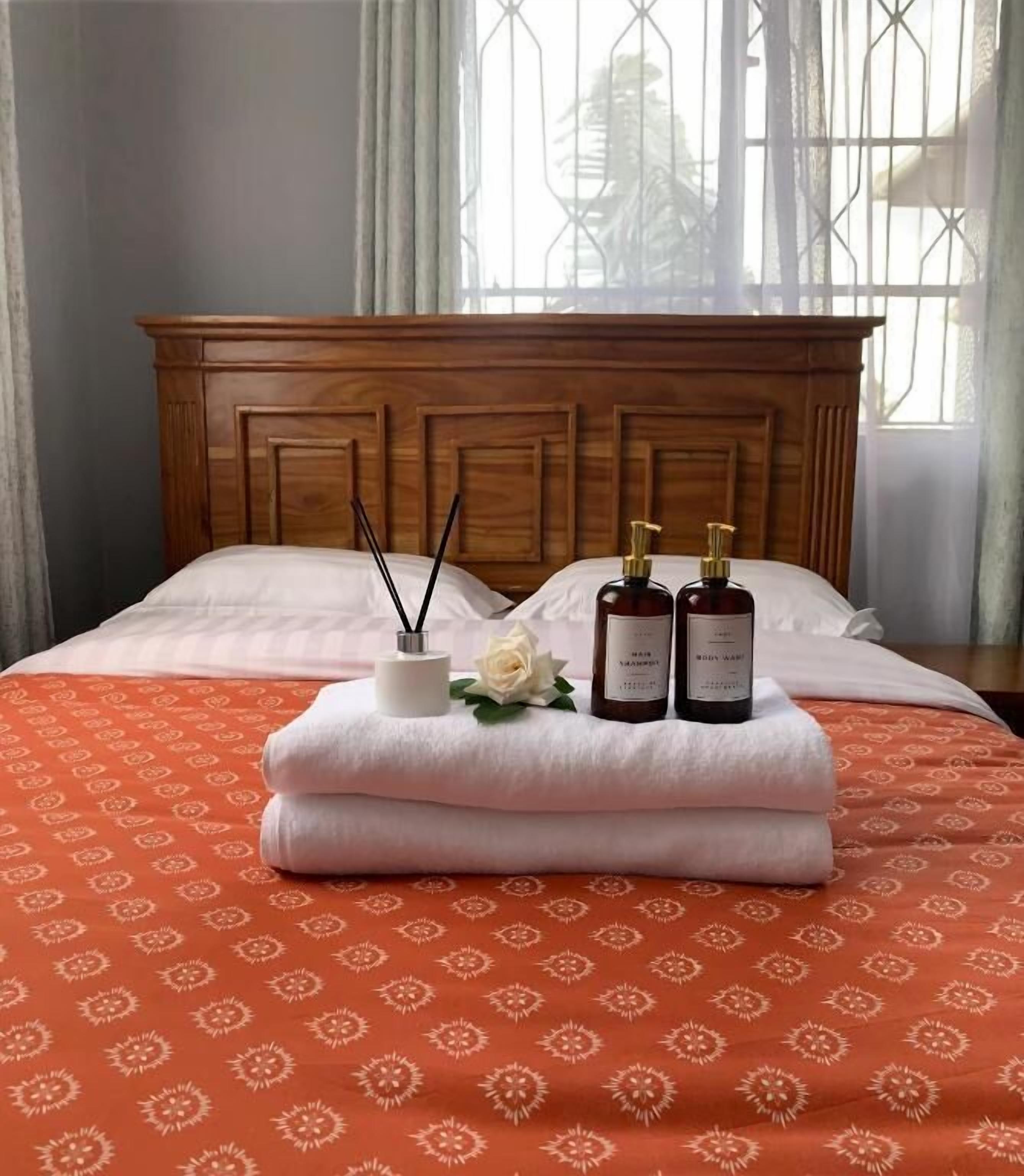 A bed with a wooden headboard, white pillows, and an orange patterned bedspread. There are two bottles and a white container with a rose on the bed.