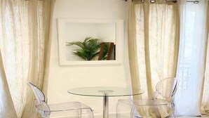 Quarto com mesa de vidro e duas cadeiras, quadro emoldurado na parede e cortinas leves.