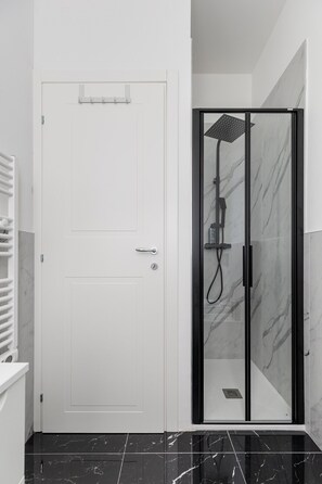 Un baño moderno con una puerta blanca, cabina de ducha con mampara de vidrio y ducha de mano, y piso de mármol.