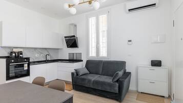 Een moderne keuken met een inbouwoven, een eethoek met een tafel en stoelen, een grijze bank en een witte kast.