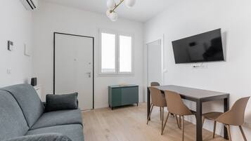 Ein modernes Wohnzimmer mit einer Couch, einem Esstisch mit Stühlen, einem an der Wand befestigten Fernseher und einer Tür zu einem anderen Raum.