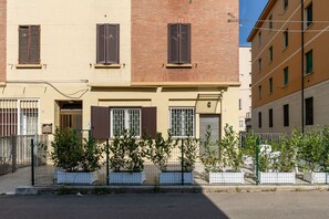 Ein zweistöckiges Gebäude mit beiger Fassade und braunen Fensterläden, das ein befestigtes Eingangstor und eine kleine Gartenanlage mit Blumentöpfen zeigt.