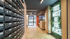 A hallway with a row of metal storage lockers on the left and a reception desk with a green wall and a neon sign on the right. - Student Only Newarke Point (Leicester)