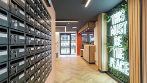 A hallway with a row of metal storage lockers on the left and a reception desk with a green wall and a neon sign on the right.