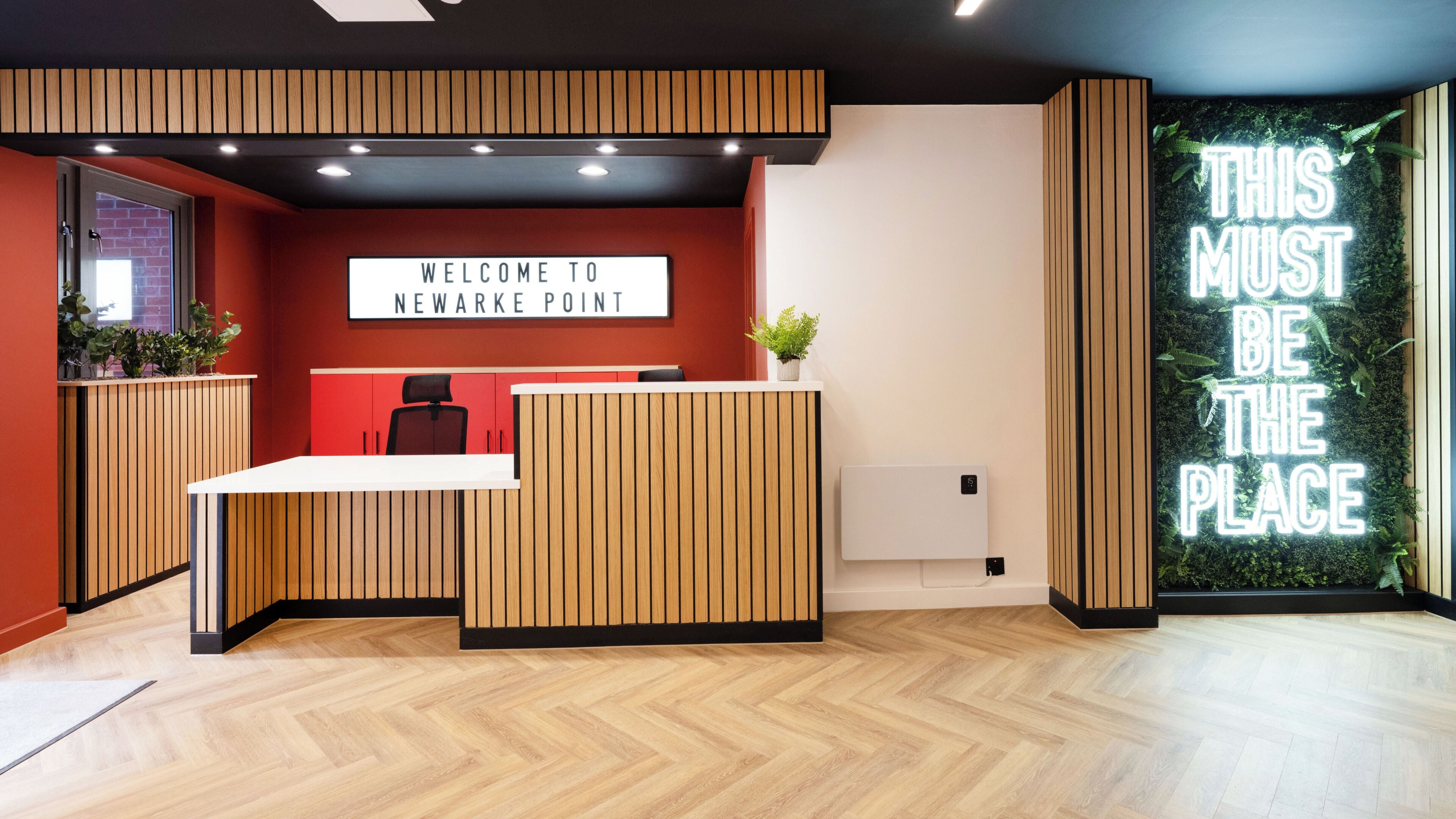 A reception desk with a 'Welcome to Newark Point' sign, a potted plant, and a neon sign stating 'THIS MUST BE THE PLACE'.