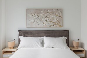 Ein Schlafzimmer mit einem Bett, zwei Nachttischen, einer Nachttischlampe auf jedem Tisch und einem an der Wand hängenden Bildzweig mit Blumen.
