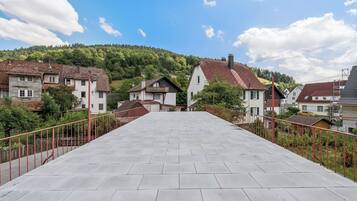 Ein gepflasterter Weg führt zu einem Wohngebiet mit Häusern, ein bewaldeter Hügel im Hintergrund und ein klarer Himmel.