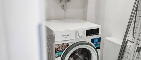 A white Siemens washing machine in a laundry room with a metal rack and a white wall.