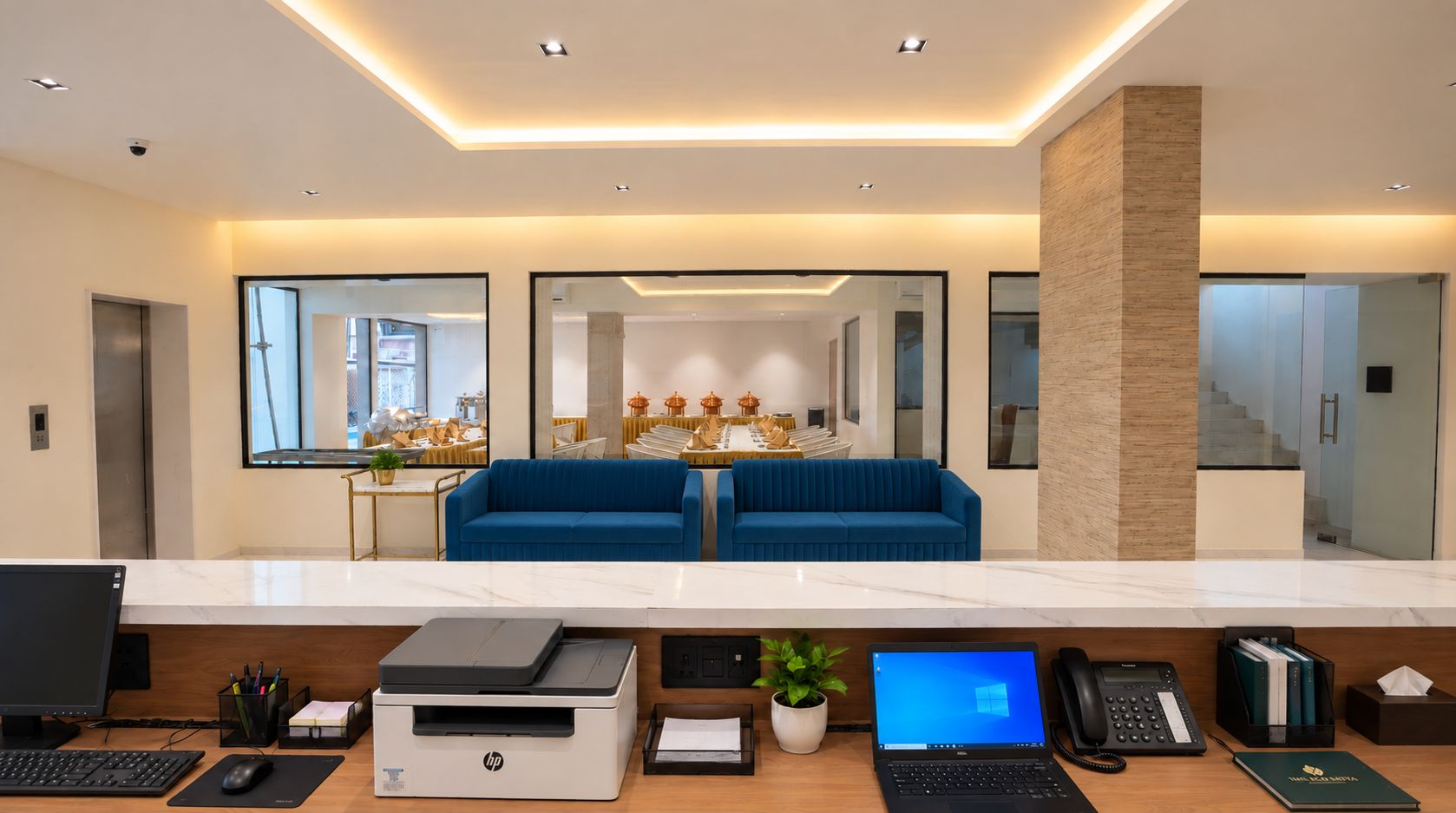 Una moderna zona de recepción con mostrador de mármol, computadoras HP y Dell, una impresora y un sofá azul.