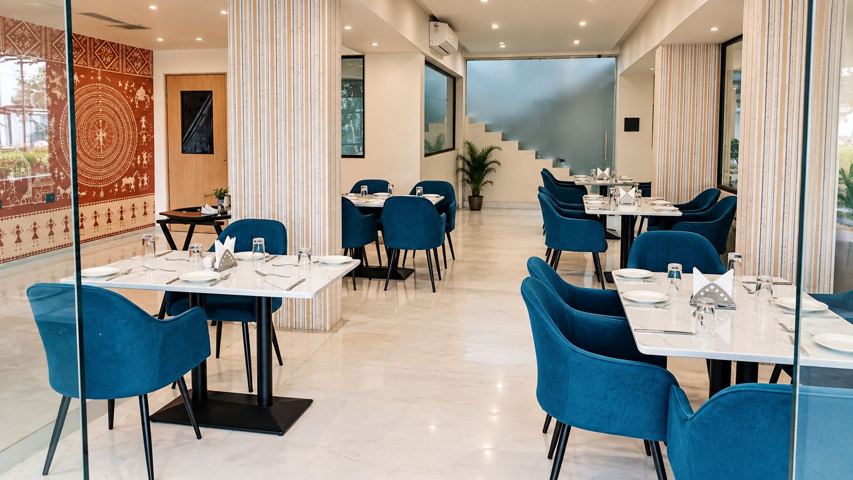 Comedor de hotel con varias mesas y sillas, una escalera al fondo y una pared decorativa con diseños.