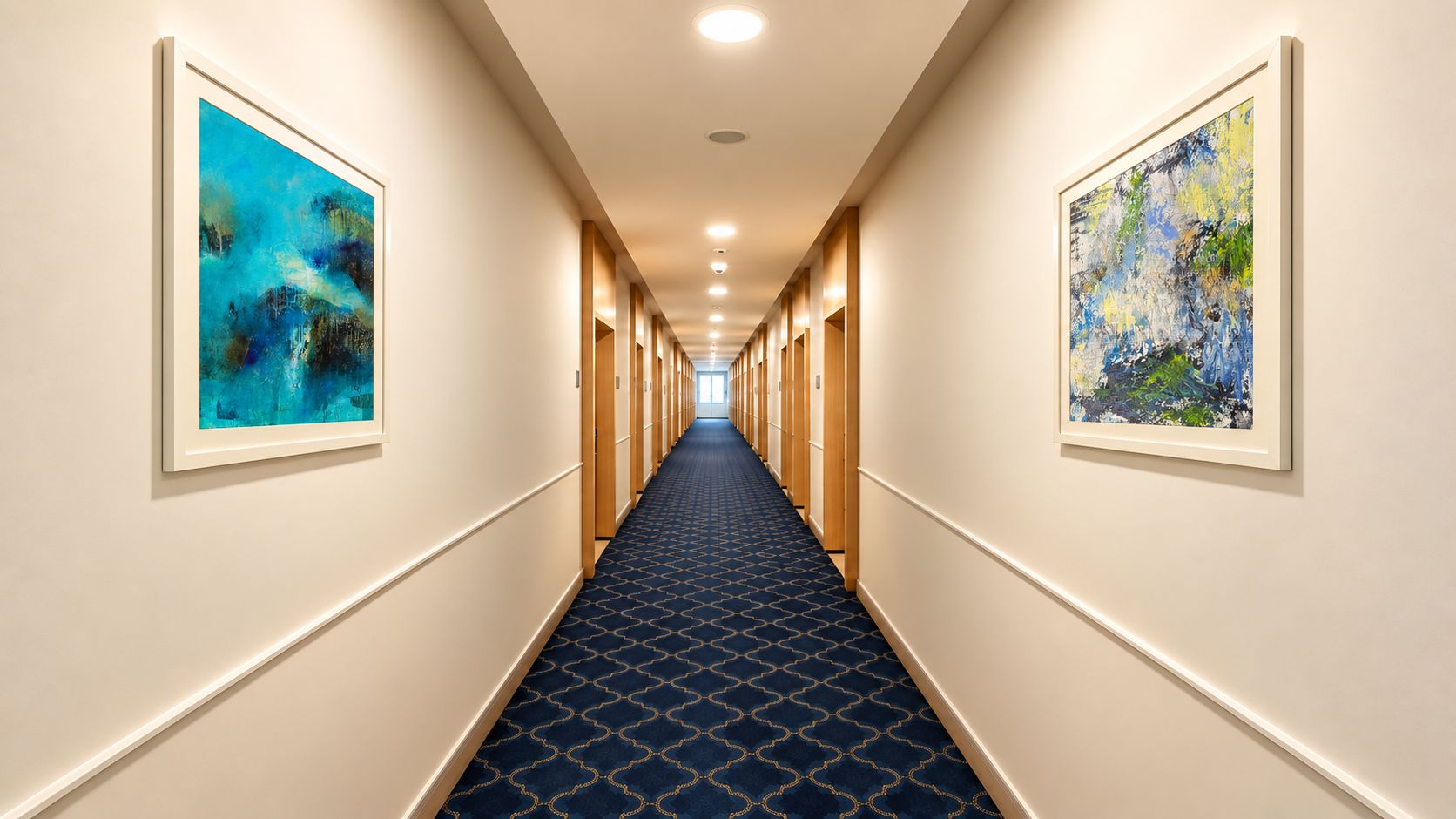 Un pasillo de hotel con cuadros abstractos enmarcados en las paredes, una alfombra estampada y varias puertas que dan a las habitaciones.