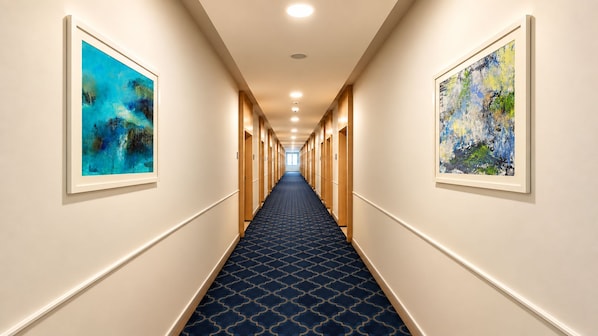 Un pasillo de hotel con cuadros abstractos enmarcados en las paredes, una alfombra estampada y varias puertas que dan a las habitaciones.