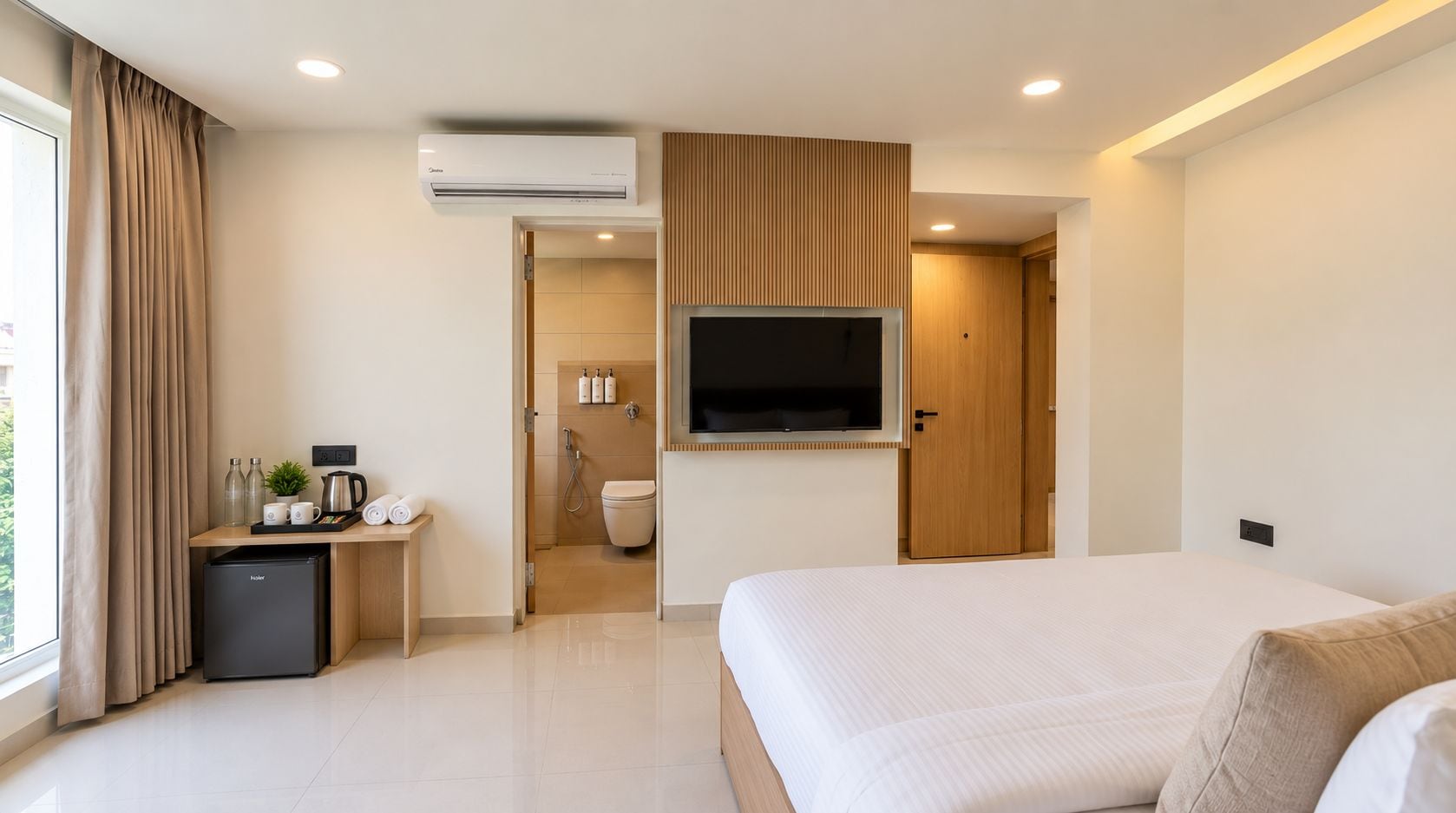 A hotel room with a bed, a desk with a coffee maker, a mini-fridge, a TV, and a bathroom visible through an open door.