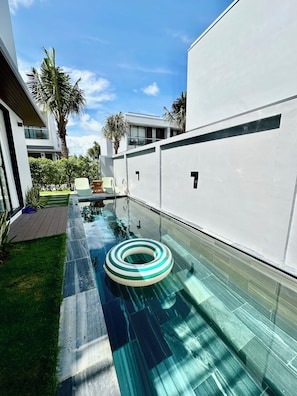 Ein moderner Außenpool mit transparentem Glasboden, umgeben von einer weißen Wand und einer Holzterrasse.