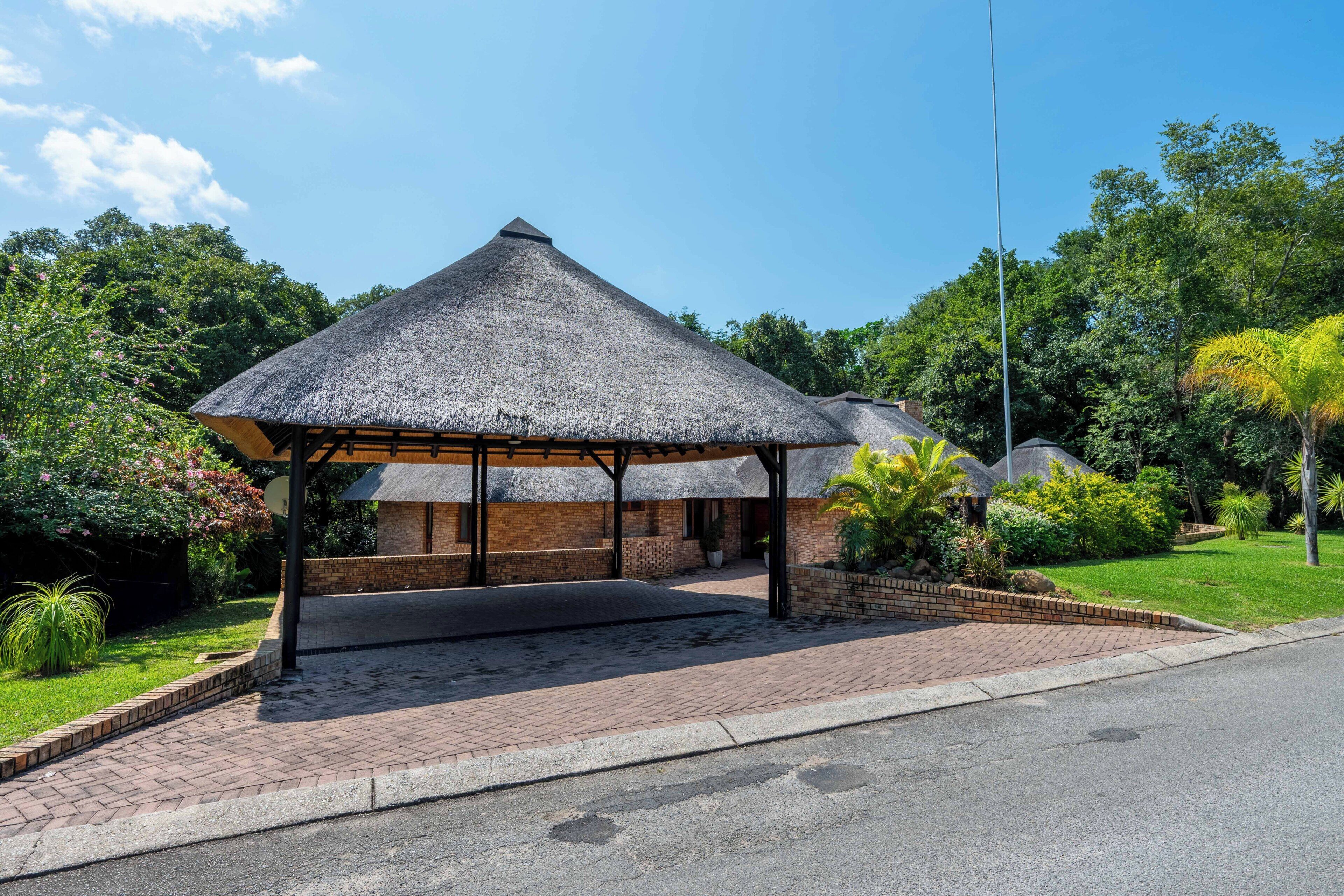 A thatched-roof structure with a brick base, surrounded by greenery and a paved walkway.