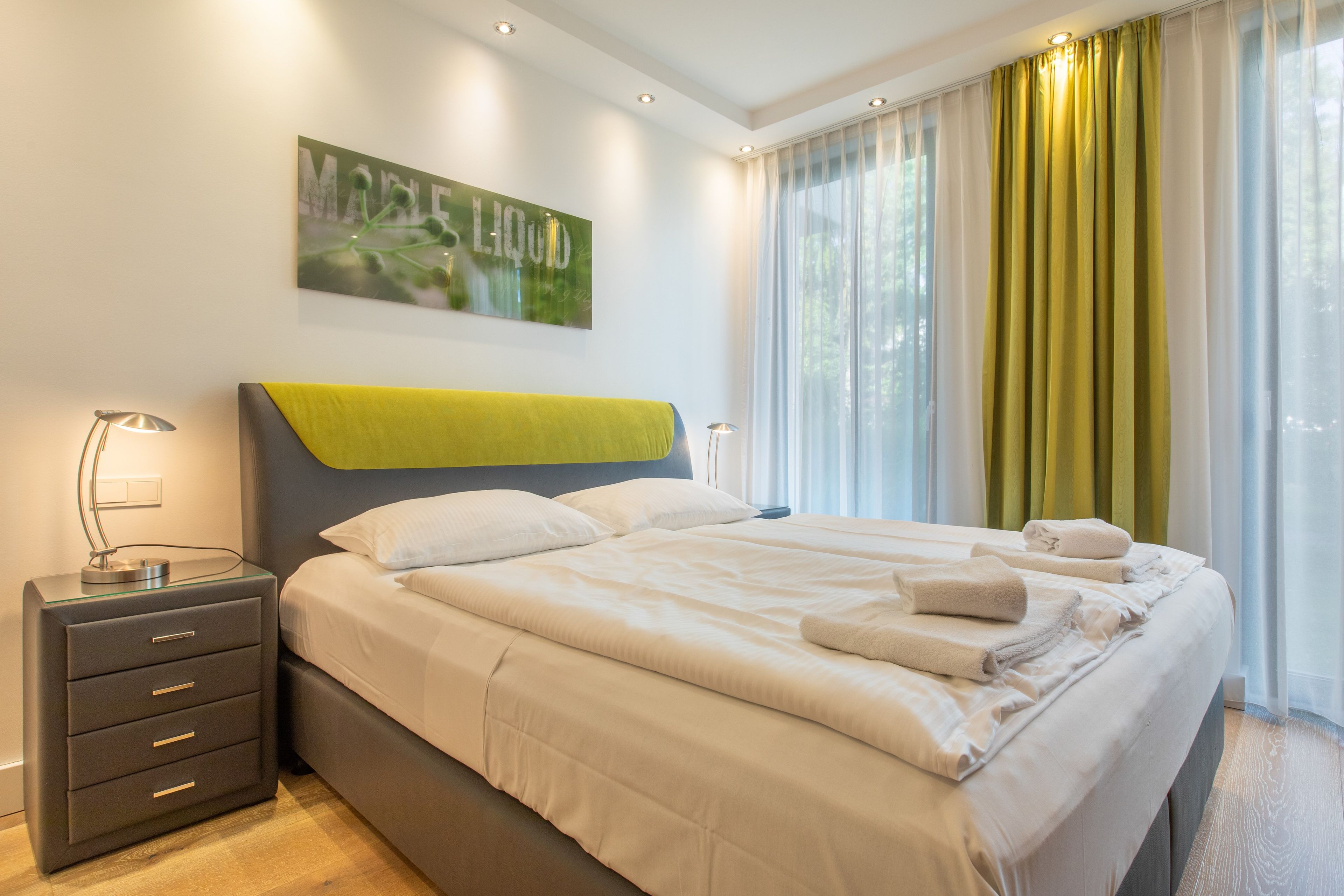 A modern hotel room with a large bed, a bedside table with a lamp, and a painting on the wall.